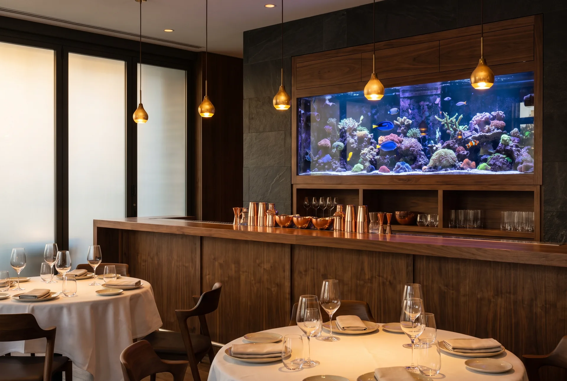 An empty upscale restaurant in the late afternoon: a large saltwater reef aquarium glows in the wall behind a long walnut bar with copper cups and glassware, tables laid with white cloths in front.